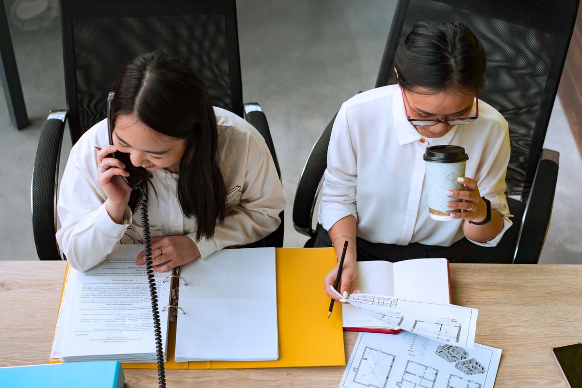 Two real estate professionals reviewing floor plans and documents at a desk with phone and notebook
